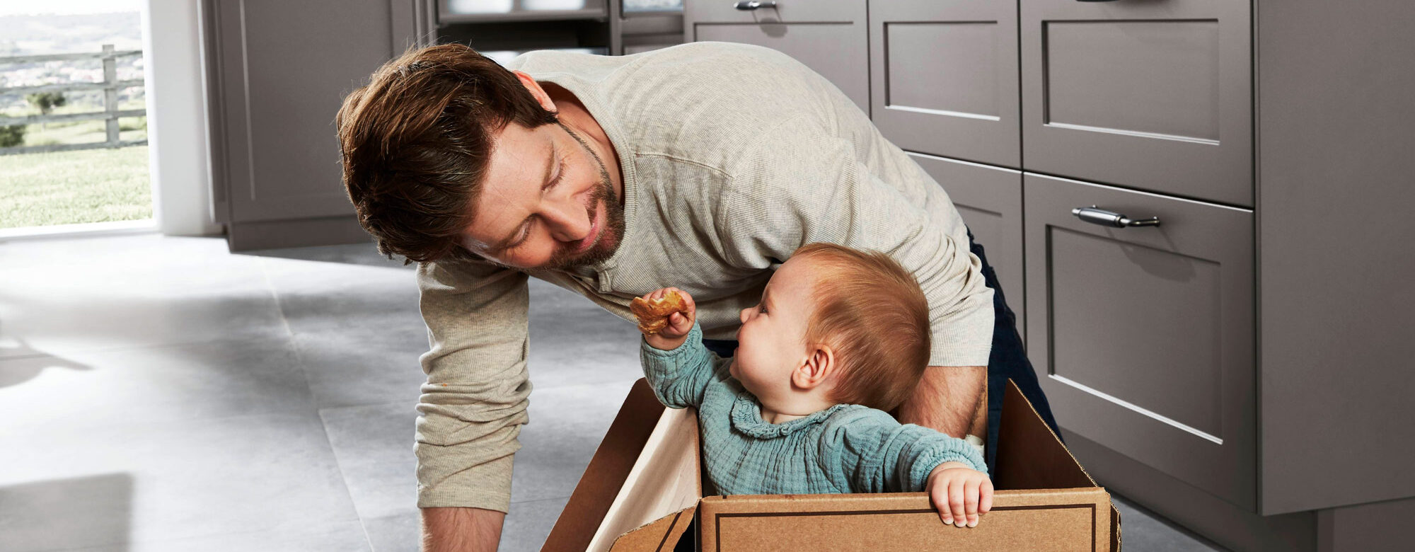Ein Mann lächelt ein Baby an, das in einem Karton in einer modernen Küche sitzt. Das Baby hält etwas zu essen in der Hand und schaut zu dem Mann auf. Die Küche besticht durch clevere Stauraumlösungen mit grauen Schränken und einer hellen, offenen Atmosphäre.