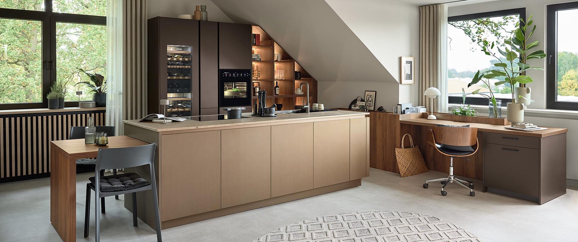 Eine moderne Kombination aus Küche und Homeoffice mit Holzschränken, einem Einbaubackofen, einem Schreibtisch, einem Stuhl, Regalen, Zimmerpflanzen, großen Fenstern, neutralen Farben und einem gemusterten runden Teppich auf hellem Fliesenboden – ein Beispiel für durchdachte Küchenplanung, die Stil und Funktionalität in einem einladenden Raum optimal vereint.